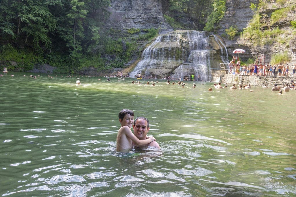Swimming at Robert H. Tremon State Park. The water is surrounded by tall stone walls covered in green foliage with a waterfall in the background.