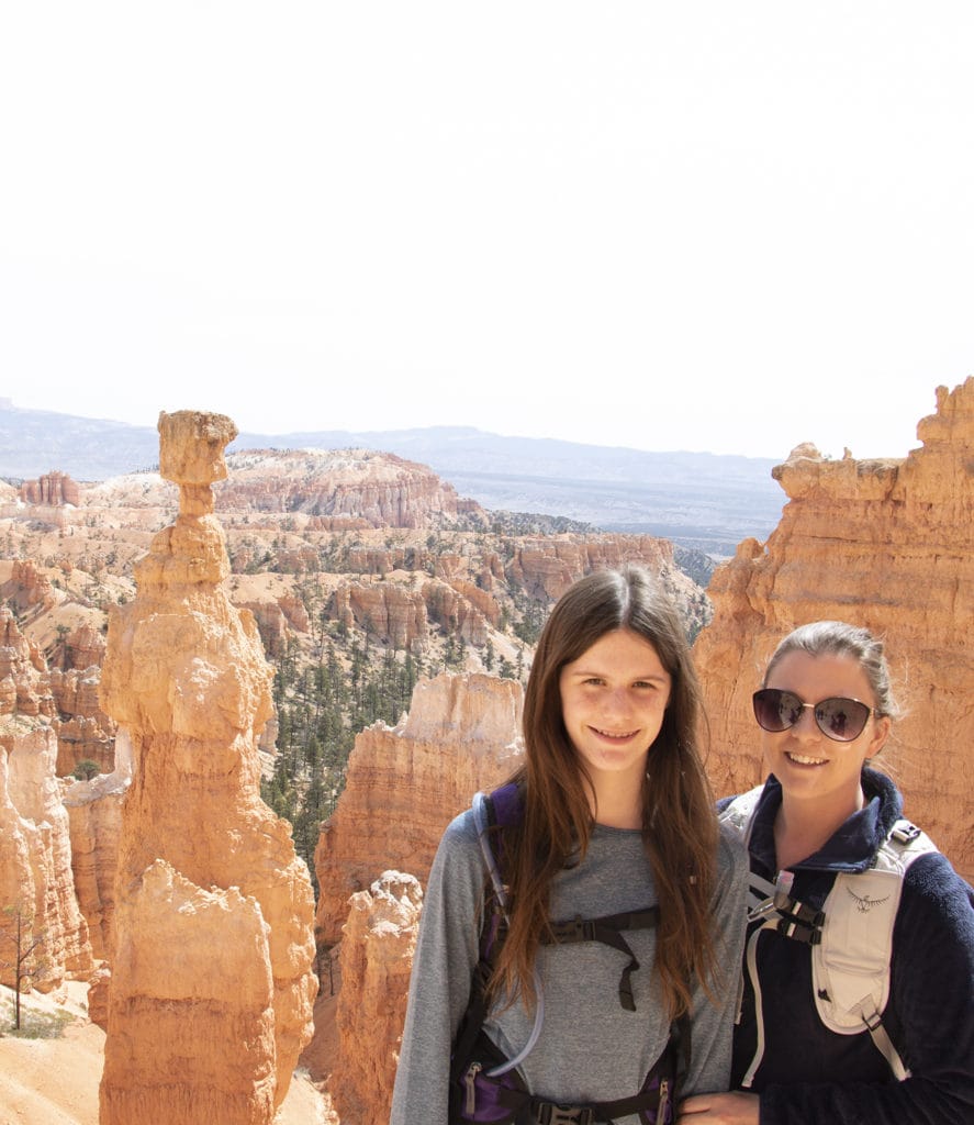 Mother and daughter standing in awe in front of Thor's Hammer, a prominent rock formation in Bryce Canyon National Park. The two figures are admiring the towering red hoodoo, showcasing the park's stunning geological wonders and the sense of wonder it inspires.