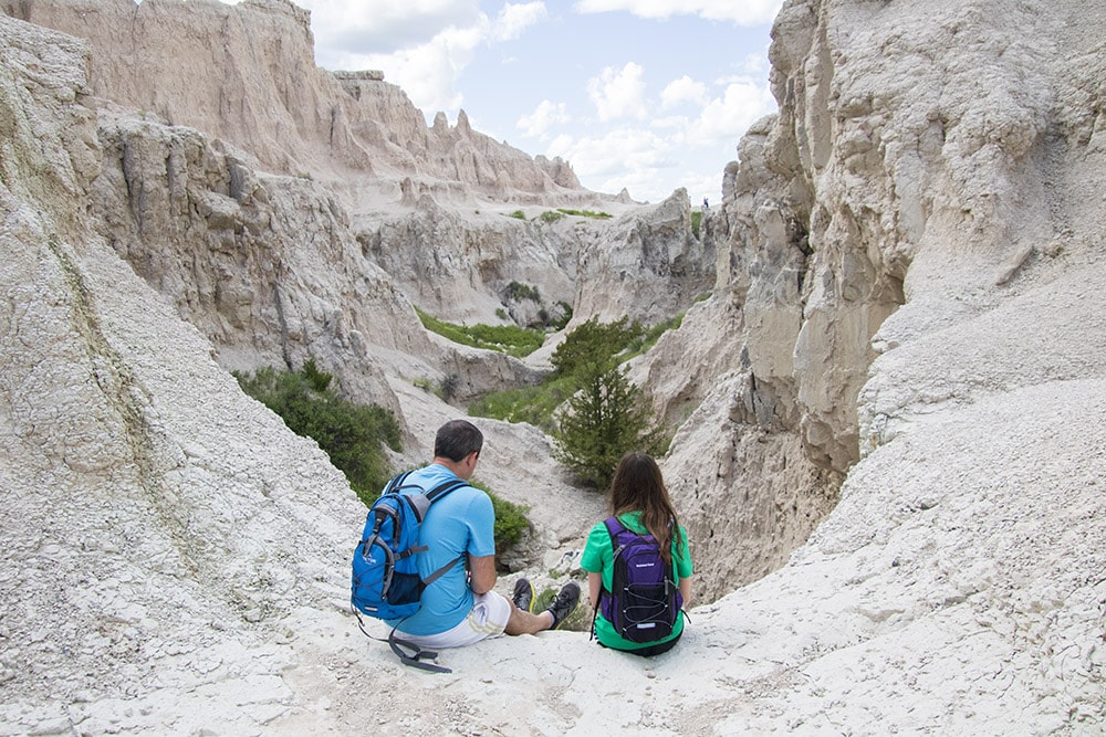 Dad and daughter enjoying the panoramic view of the Badlands from a hiking trail. They sit together, admiring the unique rock formations and rugged terrain. The image captures the bond between them and the vast beauty of Badlands National Park.
