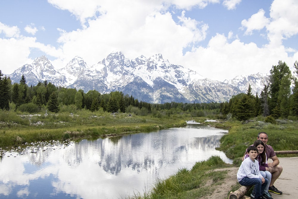 Family hiking in Grand Teton National Park sitting on a log in front of a river that shows the reflection of the snow-capped mountains.