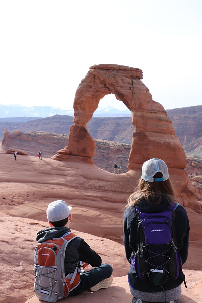 Excited kids sitting in awe in front of Delicate Arch, a magnificent sandstone arch in Arches National Park. The children are marveling at the iconic natural wonder, framed by the vivid orange rock formations, showcasing the park's captivating geology and sense of wonder.