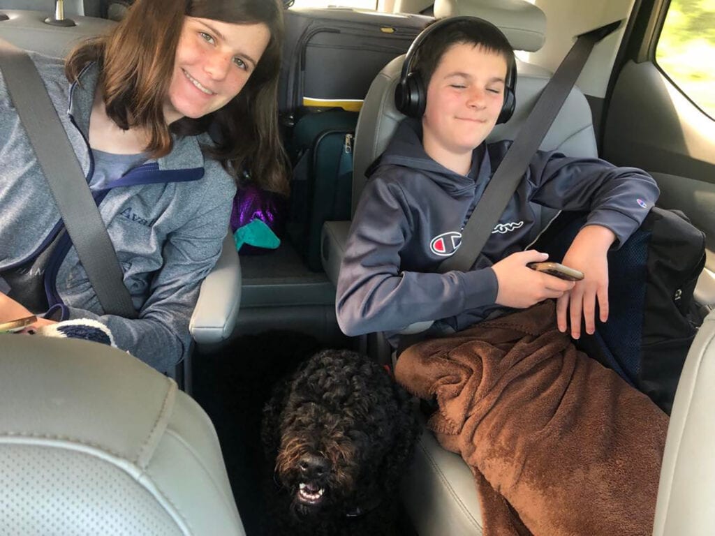This image shows a boy and a girl sitting in the back seat of a car with a dog. The boy has headphones on and there is luggage behind the kids.