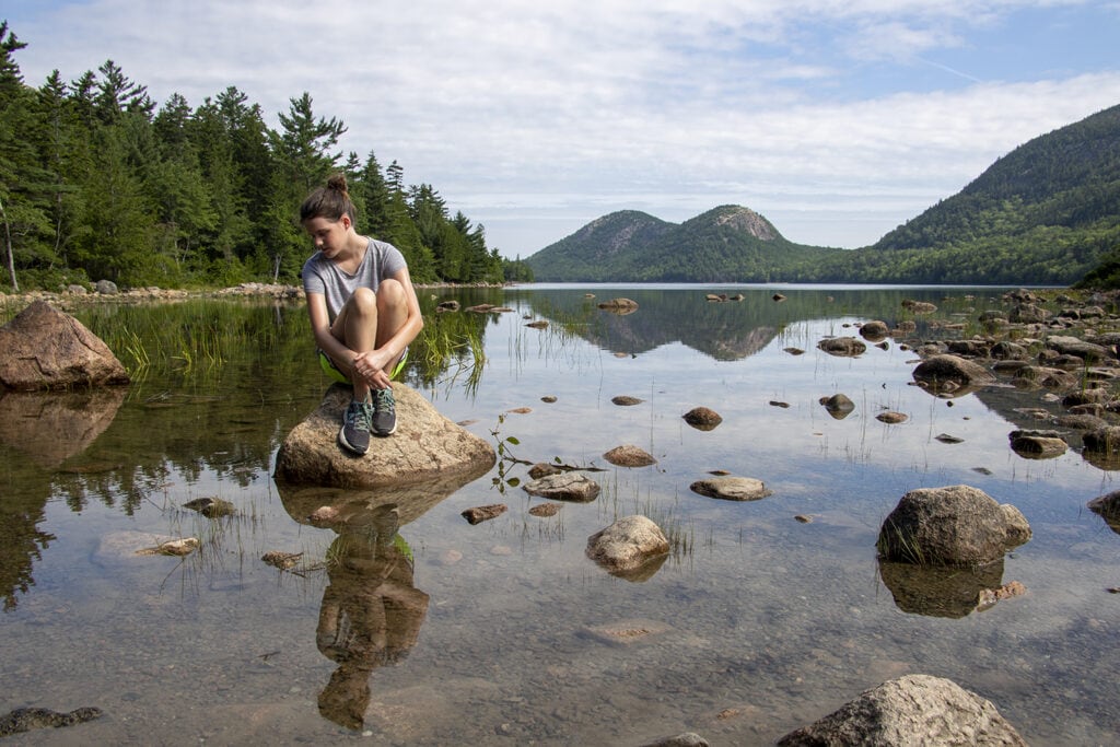 A girl sitting on a large rock in the water at Jordan Pond in Acadia. You can see her reflection in the clear shallow water.