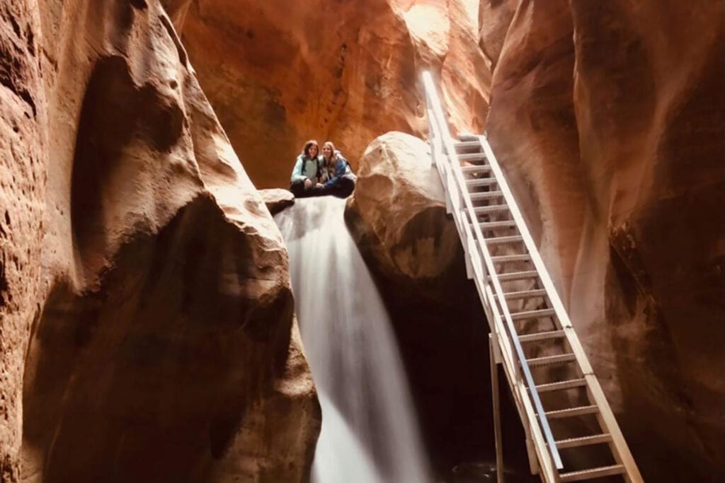 A beautiful waterfall with crystal-clear water cascading down over rocks in a narrow slot canyon. A silver metal staircase leans against the canyon wall near the waterfall, leading to the upper portion of the falls. Sunlight streams in from above, casting a warm glow on the water and surrounding canyon walls.