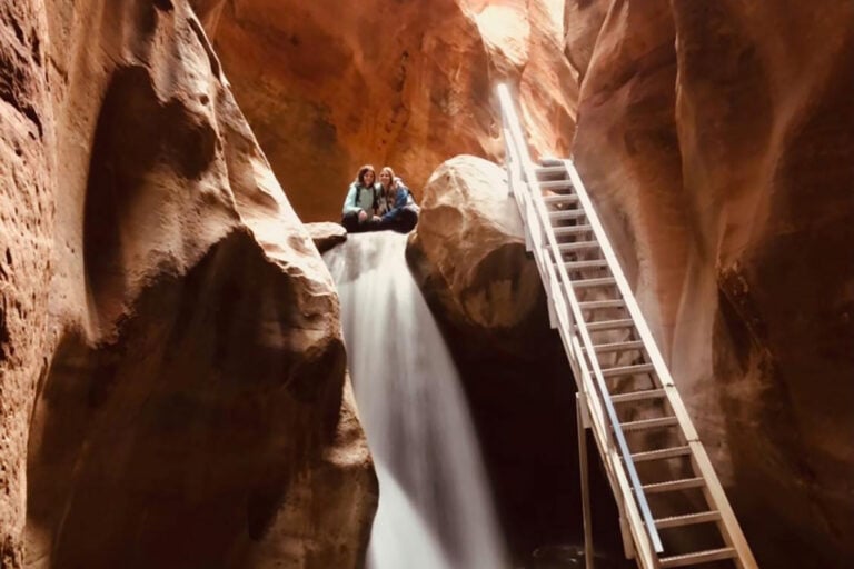 Beautiful Kanarra Falls with crystal-clear water cascading down over rocks in a narrow slot canyon. A silver metal staircase leans against the canyon wall near the waterfall, leading to the upper portion of the falls. Sunlight streams in from above, casting a warm glow on the water and surrounding canyon walls.