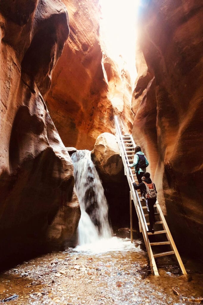 Two people climbing up the metal staircase at the first waterfall inside of the Kanarra Falls slot canyon. The ladder sits right next to the waterfall.