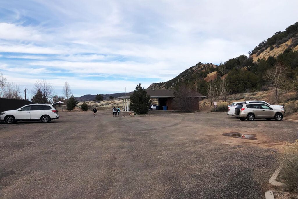 A view of the Kanarra Falls parking lot, showing a large paved area with several cars parked and a small building on the right-hand side. There are also picnic tables and restrooms in or by the brick building pictured and a shoe-washing station near the entrance and behind the building.