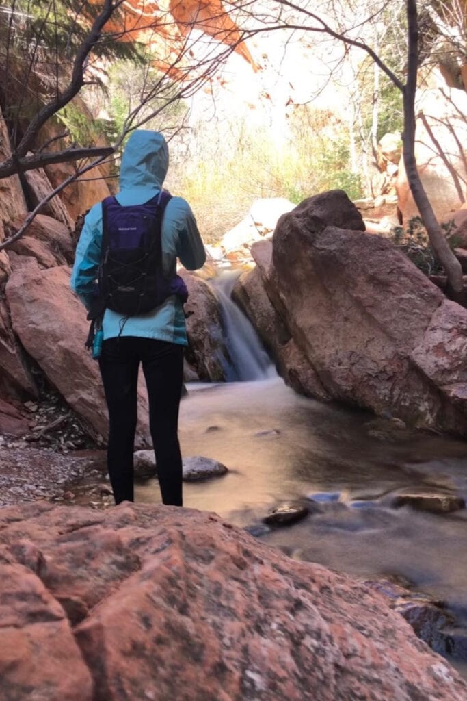 A girl looking at a small waterfall on the way to Kanarra Falls slot canyon. The waterfall drapes over large red boulders.