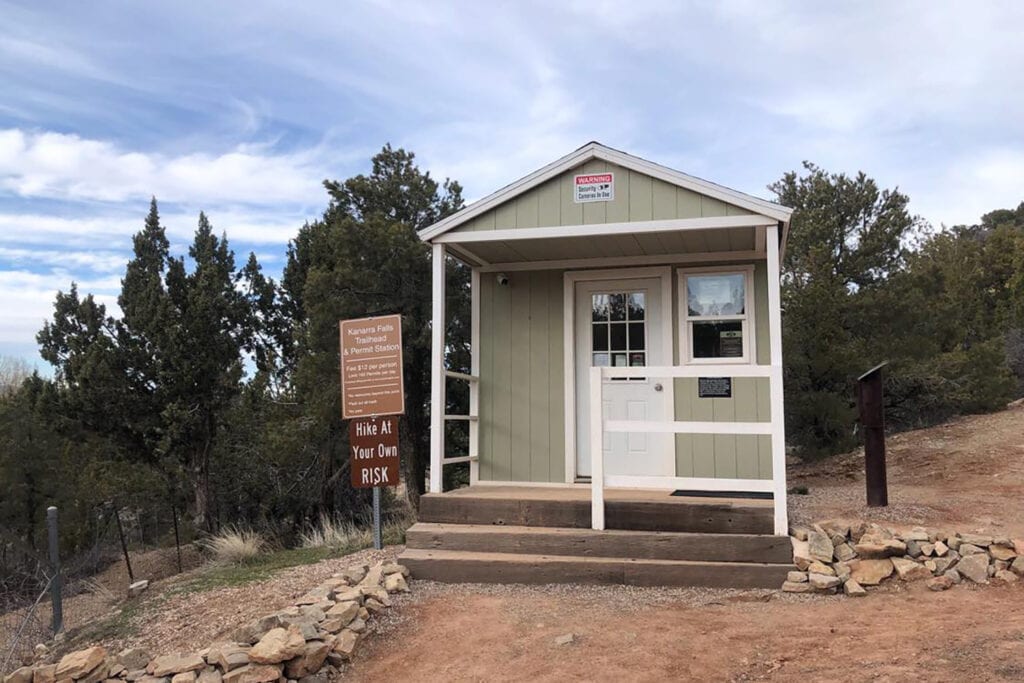 A photo of the permit station at Kanarra Falls, showing a sign with instructions for obtaining hiking permits. The station is a small wooden structure with a sloping roof, door and window. The window is where you show your permit and get waiver papers to fill out. The surrounding area features rocky terrain with large trees.