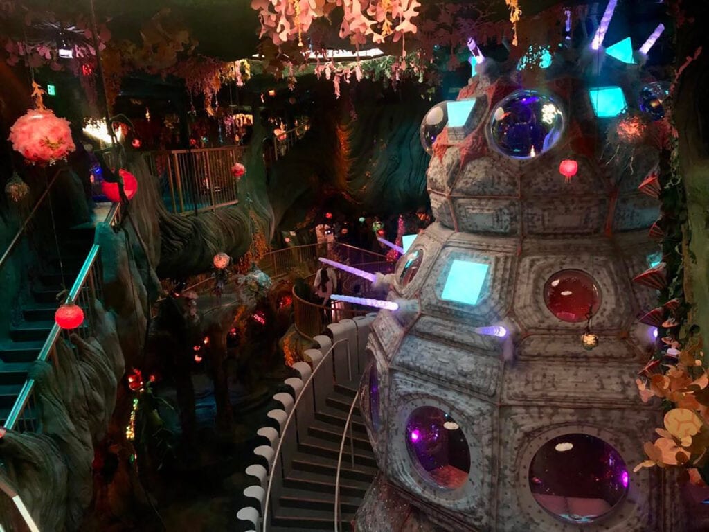 This image shows part of the treehouse room at meow wolf denver. The treehouse room has an elevated wooden structure with multiple levels and platforms, adorned with greenery and surrounded by intricate patterns and colorful lights. The room features a variety of furniture pieces and quirky decor, including a couch swing, a bathtub, and a telescope. The focal point is a towering tree-like structure, complete with branches and foliage, that extends from the ceiling to the floor.