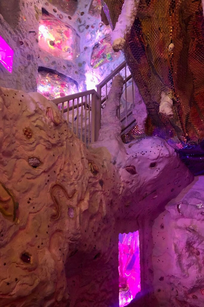 Altar-like structure in a dimly lit room with pink walls and floors at Meow Wolf Denver. The centerpiece of the room is a round table with candles, vases, and sculptures, surrounded by plush pink chairs. Various art installations and decor elements can be seen on the walls, including a large painting, a framed photo, and hanging pink tassels. The overall vibe of the room is mysterious and ethereal.