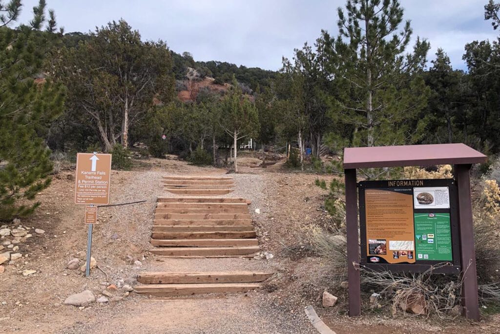 A view of the wooden stairs leading to the Kanarra Falls trailhead, showing a sturdy staircase built into a rocky hillside. The stairs have some landings leading up to a dirt path. The surrounding landscape features large evergreen trees.