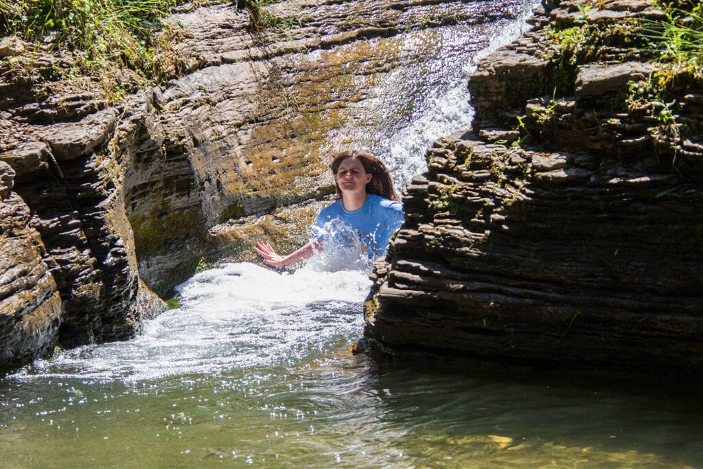 Person joyfully sliding down the natural waterslide at devil's bathtub sd trail, Spearfish Canyon, surrounded by scenic beauty and rock formations.