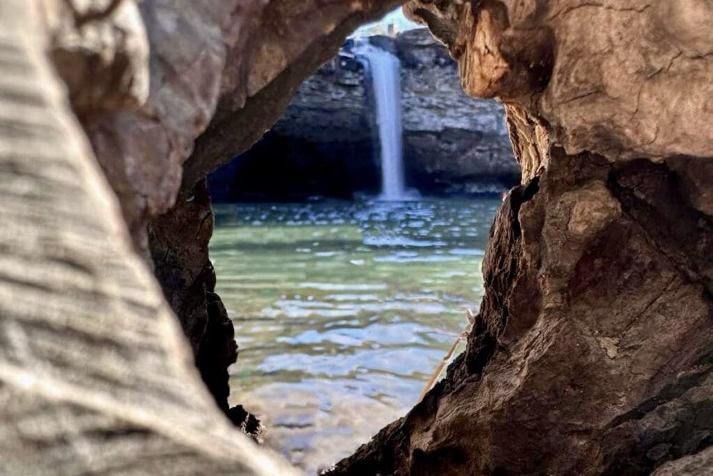 View from the base of DeSoto Falls, framed by a natural wooden aperture. The waterfall cascades gracefully, surrounded by lush greenery. The perspective is captured through a unique opening in a wooden structure, adding a natural frame to the scenic beauty.