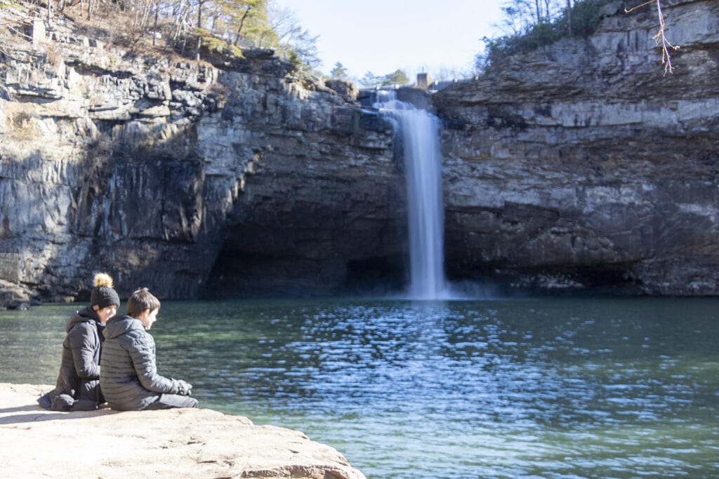 Relaxing at the base of DeSoto Falls, surrounded by nature's tranquility. The gentle sounds of cascading water provide a soothing backdrop as visitors enjoy the serene atmosphere. Lush greenery frames the scene, creating a peaceful spot for relaxation by the waterfall.